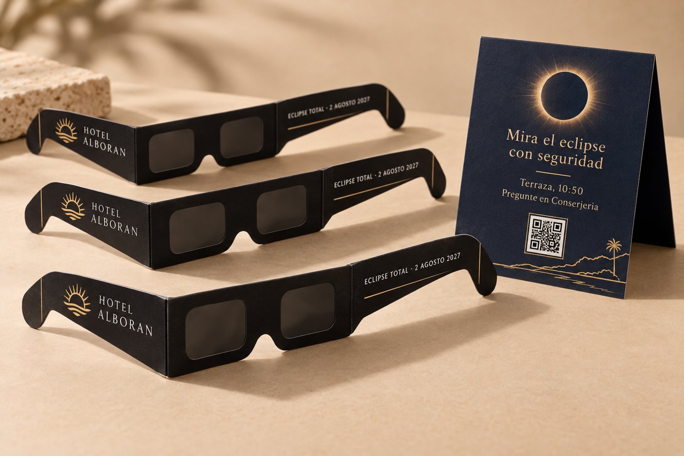 Three black eclipse glasses printed with a hotel logo and 'Eclipse total 2 agosto 2027', next to a navy fold-out card with a QR code